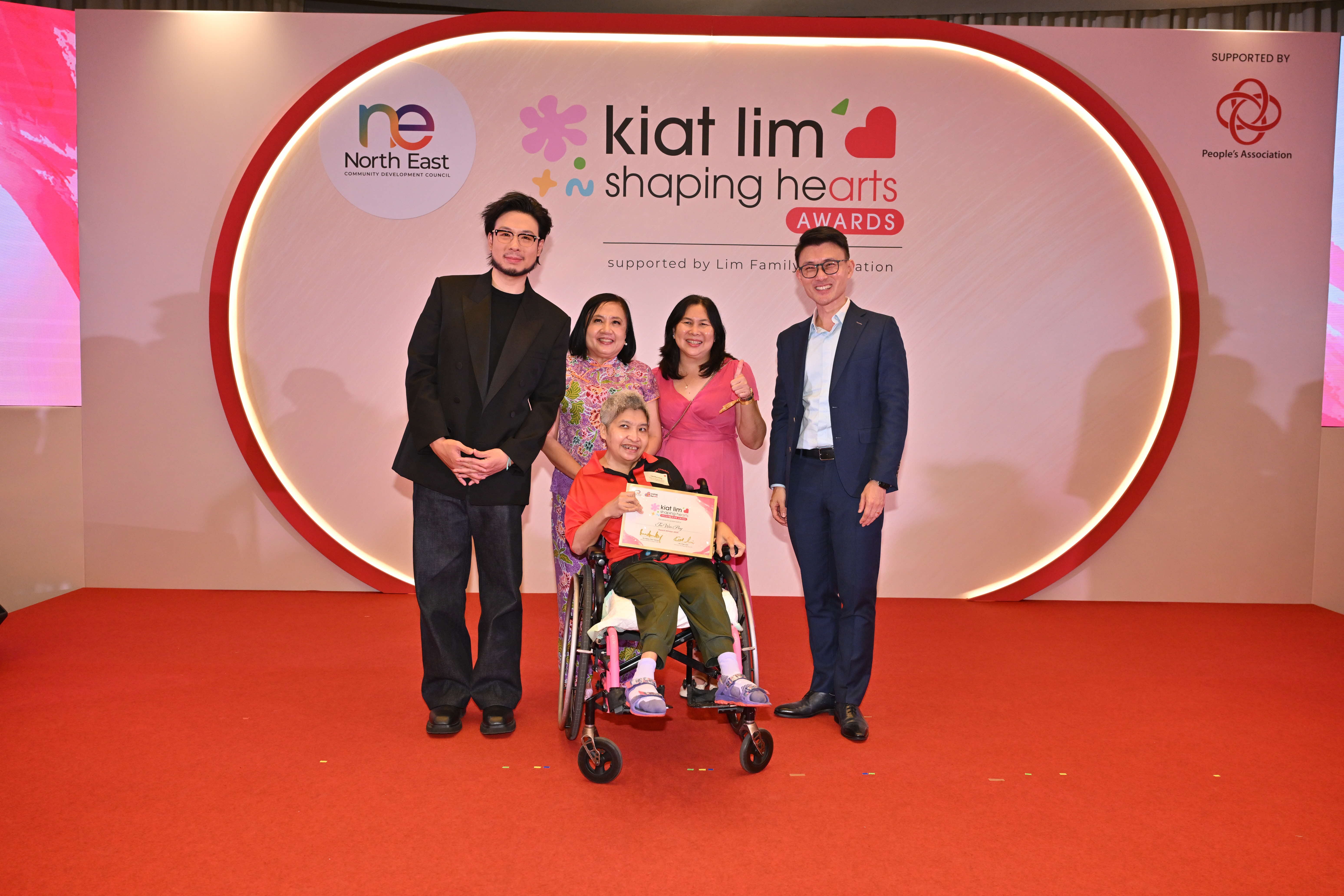 Award recipient standing on stage with the presenter, holding their certificate and posing for a group photo during the award ceremony, with a backdrop reading ‘Kiat Lim-Shaping Hearts Awards'.